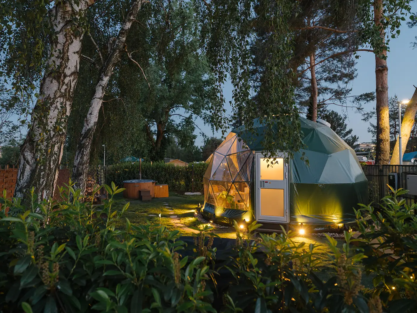 Geodesic dome lit at dusk among birch trees, with a wood-fired hot tub in the garden